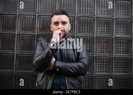 Hispanic gay man in leather jacket with finger on his nose standing against glass wall outdoors Stock Photo