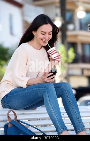 Smiling Mixed race woman drinking takeaway beverage while using smartphone sitting on bench in park during daytime Stock Photo
