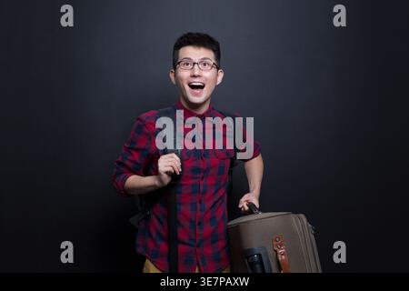 Cheerful Chinese man in eyeglasses carrying backpack and luggage looking away while standing against dark background in studio Stock Photo