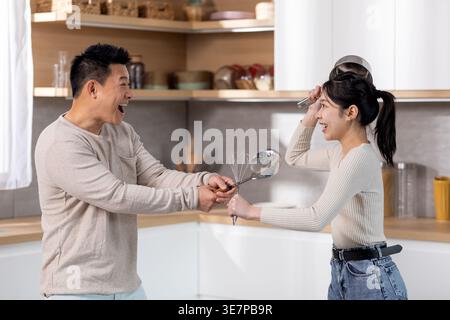 Cheerful Asian couple having fun holding kitchen utensils and making fight while standing in kitchen at home Stock Photo