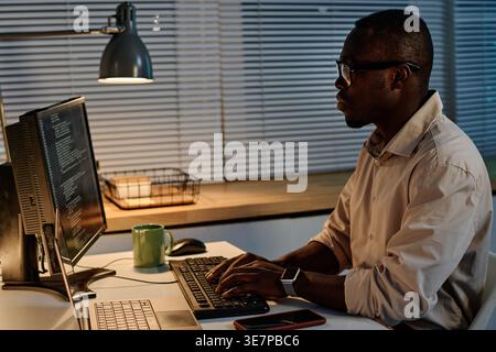 Focused Black male programmer in eyeglasses examining computer codes working overtime in office Stock Photo