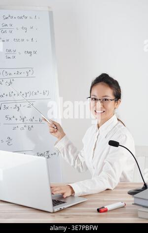 Smiling Asian female math teacher sitting at her desk with microphone and laptop explaining formula to students in classroom Stock Photo
