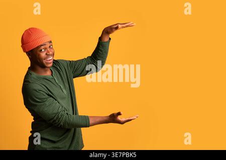 Black man in beanie holding invisible object while standing against yellow background in studio Stock Photo