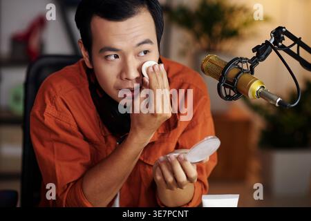 Asian male online seller applying compact foundation on his face sitting at table streaming live from home studio Stock Photo