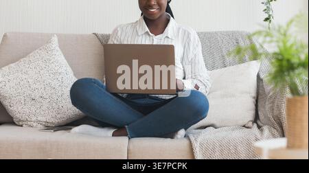 Cropped image of Black woman with twists working on laptop sitting cross-legged on couch in the living room at home Stock Photo