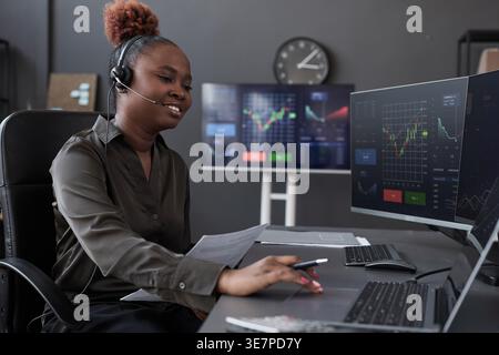 Black female trader with high bun wearing headset talking to customer and examining financial graphs using laptop while sitting at desk in office Stock Photo