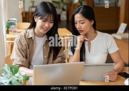 Asian female friends working remotely together using laptop and tablet in coffee shop Stock Photo