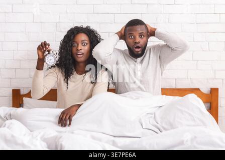 Black man with hands behind head sitting on bed with his wife holding alar clock in bedroom at home Stock Photo