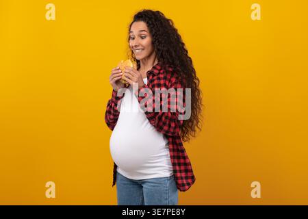 Smiling Middle Eastern pregnant woman with curly hair holding and looking at burger while standing against yellow background in studio Stock Photo