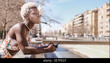 Serious Black non-binary person looking away leaning on bridge railings in park on sunny day Stock Photo