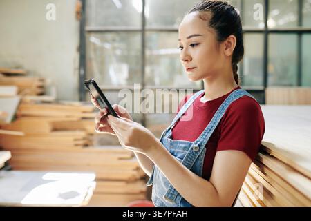 Asian woman checking text messages on her smartphone standing in carpentry woodshop Stock Photo