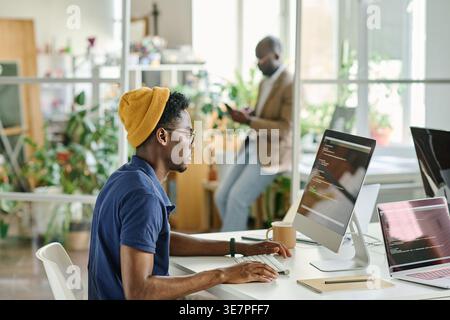 Black male programmer wearing yellow beanie and eyeglasses working on computer with codes while sitting at desk in office Stock Photo