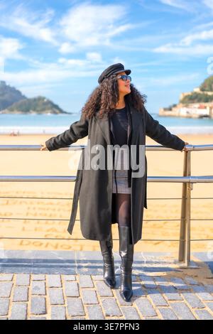 Hispanic woman with curly hair looking away leaning against railingss on beach Stock Photo