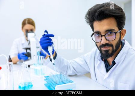Indian male scientist working with chemical liquids in flask near caucasian female colleague using microscope in laboratory Stock Photo