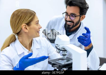 Smiling Indian male and caucasian female scientists wearing lab coats and latex gloves using microscope doing sample analysis together in laboratory Stock Photo