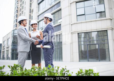 Smiling Chinese male and female architects wearing white hard hats shaking hands after closing business deal while standing on city street Stock Photo