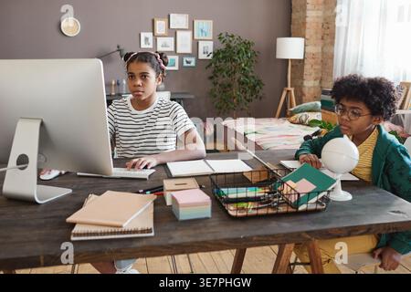 Focused Black boy with curly afro wearing eyeglasses sitting at table with his sister using tablet and computer studying together at home Stock Photo