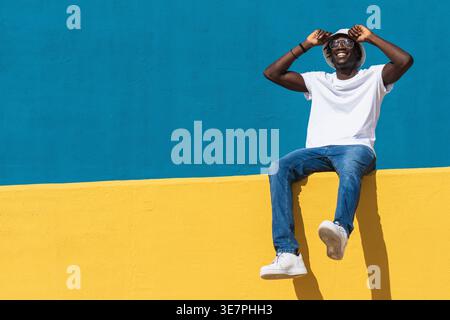 Cheerful Black man with locks wearing white shirt and jeans adjusting bucket hat sitting on yellow fence against blue wall outdoors on sunny day Stock Photo