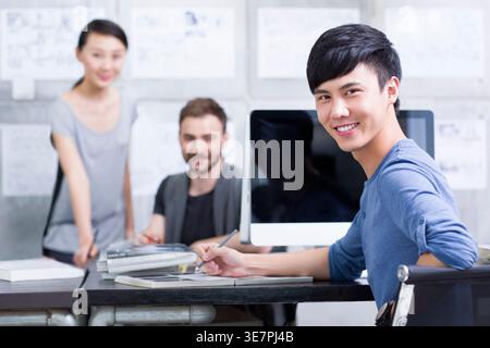 Chinese male office worker sitting at desk with computer near workmates in office Stock Photo