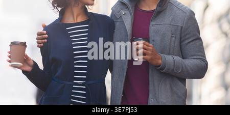Smiling Black couple looking at each other holding cup of coffee walking on city street during daytime Stock Photo