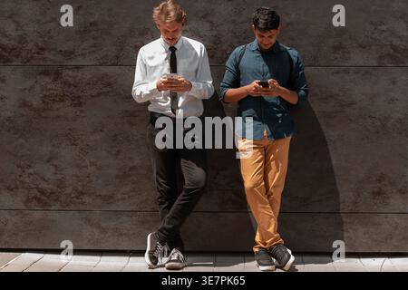 Middle Eastern and caucasian men with legs crossed using smartphones leaning against black wall outdoors on sunny day Stock Photo