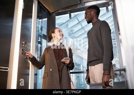 Smiling Black businessman standing and chatting with caucasian female colleague pressing button in elevator at the office Stock Photo