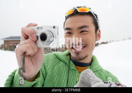 Smiling Chinese man with sunglasses on head taking picture using camera while standing in snowy street during winter Stock Photo