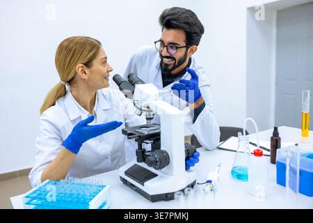 Smiling Indian male and caucasian female scientists wearing lab coats and latex gloves using microscope doing sample analysis together in laboratory Stock Photo