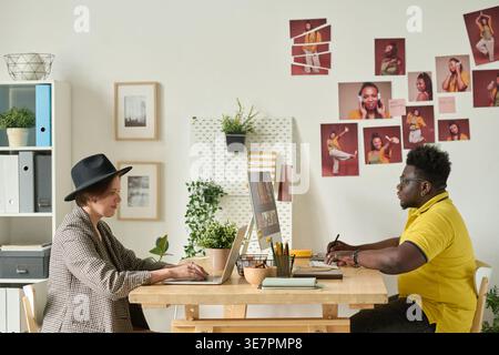 Black male editor in yellow polo shirt using graphic tablet for retouching photos on computer sitting at table with caucasian female colleague in o... Stock Photo