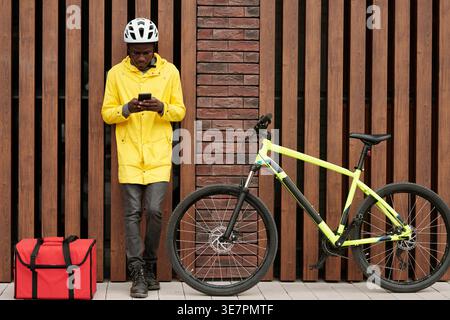 Tired Black delivery man leaning against wooden wall checking new online orders on smartphone standing on pavement near delivery food box and bicyc... Stock Photo