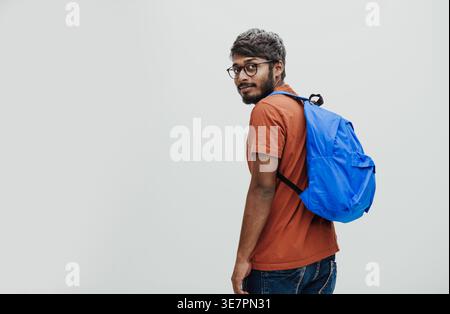 Serious Indian man in orange shirt with eyeglasses and blue backpack standing while holding notebook against white background in studio Stock Photo