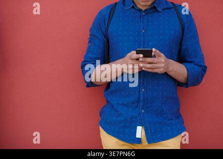 Middle Eastern man using smartphone leaning against red wall outdoors Stock Photo