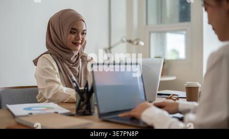 Smiling Muslim business woman wearing hijab sitting at table with female colleague working on laptops in the office Stock Photo