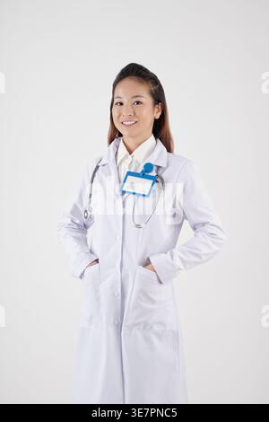 Smiling Asian female doctor wearing labcoat standing with hands in pockets against white background in studio Stock Photo