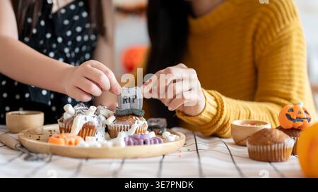 Asian mother and daughter decorating Halloween cupcakes together while sitting at table in the kitchen at home Stock Photo