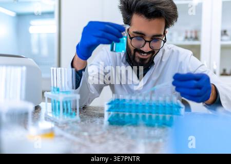 Smiling Indian male scientist wearing lab coat and latex gloves sitting at table and testing chemical sample in flask in laboratory Stock Photo
