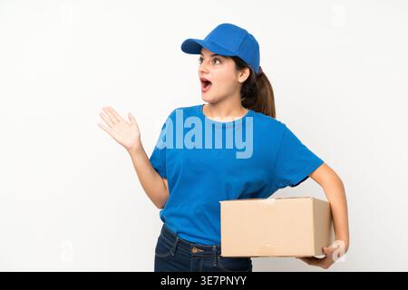 Surprised Hispanic delivery woman wearing blue cap looking away holding cardboard box standing with open hand against white background in studio Stock Photo