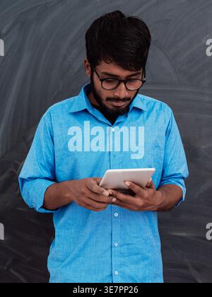 Indian man in blue long sleeves with eyeglasses using tablet while standing against chalkboard in classroom Stock Photo