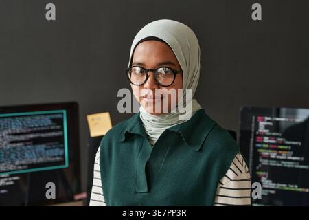 Mixed race Muslim female software developer wearing eyeglasses standing against multiple computer screens placed on table in office Stock Photo