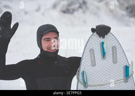 Smiling Mixed race man in surfing suit holding surf board and looking away while standing on snowy mountain during winter Stock Photo