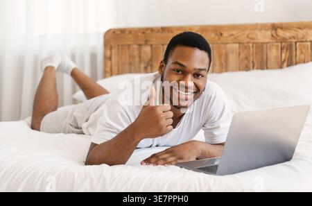 Smiling Black man in white shirt gesturing thumbs up while lying on bed with laptop at home Stock Photo