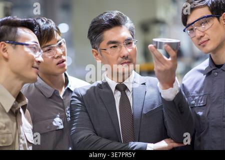 Serious Chinese businessman holding and checking machine parts while standing with male mechanical engineers in the factory Stock Photo