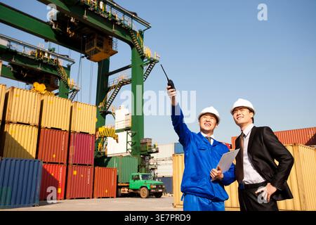 Chinese male worker pointing away standing with businessman wearing hard hat standing in shipping port during daytime Stock Photo