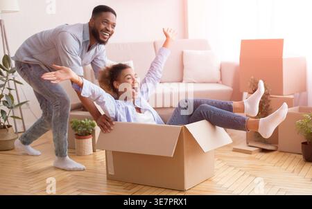 Cheerful Black man having fun pushing wife sitting in moving boxes celebrating relocation in new home Stock Photo