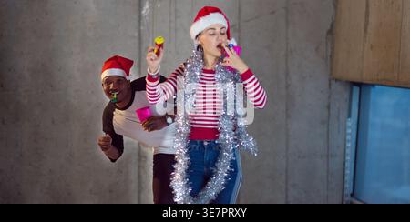 Cheerful Black man in Santa hat peeking behind caucasian female colleague celebrating New Year against gray wall in a new unfinished startup office Stock Photo