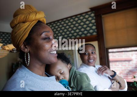 Black non-binary person standing with wife carrying their baby in the living room at home Stock Photo