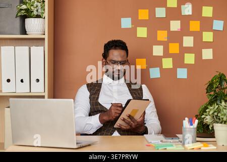 Serious Asian businessman wearing eyeglasses using tablet with stylus pen sitting at table with laptop in office Stock Photo