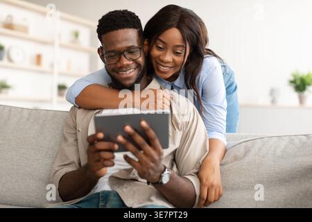 Black man using tablet sitting on sofa with wife embracing him from behind at home Stock Photo