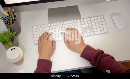 Man wearing maroon hoodie using computer while sitting at table in office Stock Photo