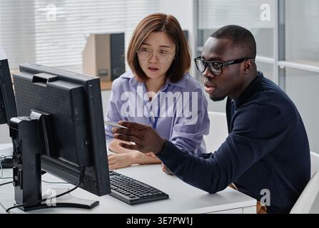 Serious Black male programmer in eyeglasses pointing at monitor and discussing presentation with Asian female colleague in office Stock Photo